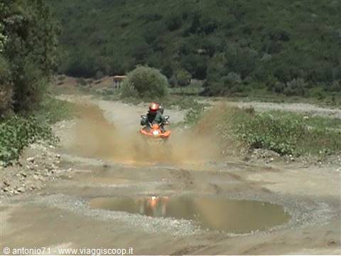 Orange quad bike splashing through a puddle on a dirt road among green hills