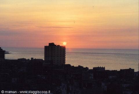 Low sun over the sea behind a tall building and city rooftops