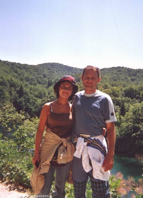 smiling couple in hat and t-shirt beside a turquoise lake surrounded by green woods