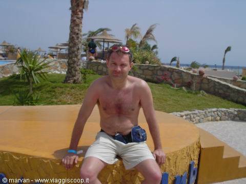Shirtless man sits on yellow structure among palms and beach umbrellas by the sea