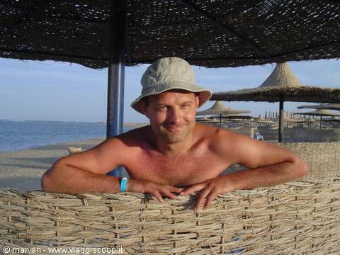 Man in hat smiling, leaning on a straw beach umbrella by the sea