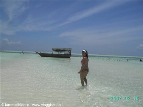 Woman in bikini wades in shallow turquoise water, wooden boat behind