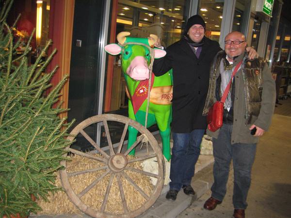 Two smiling men pose beside a green cow statue and an old wooden cart