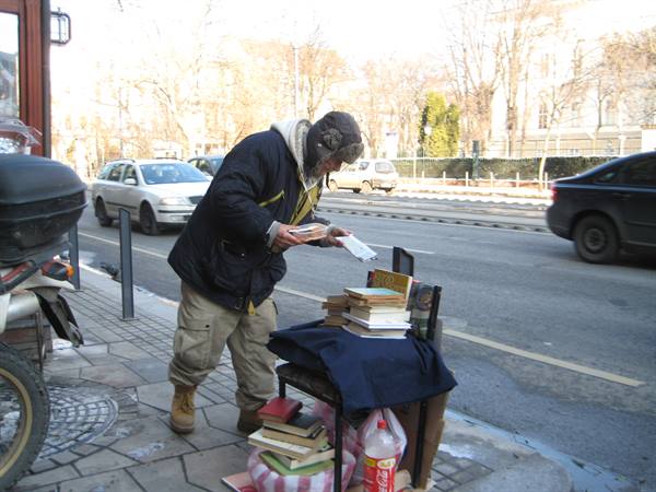 Man in fur hat selling used books from a chair on the sidewalk