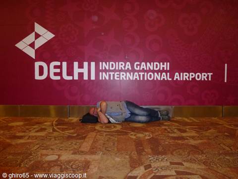 Traveler sleeping on the carpeted floor under the red Indira Gandhi International Airport sign in Delhi