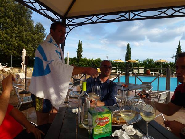 Men toast with white wine at a table beside the pool, one wrapped in a towel