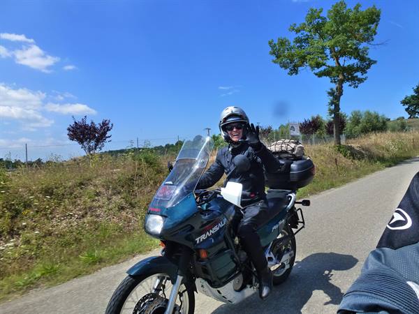 Helmeted rider waves from a green Honda Transalp on a sunny country road