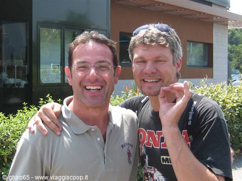 Two smiling men hug outside a building, one making the OK gesture