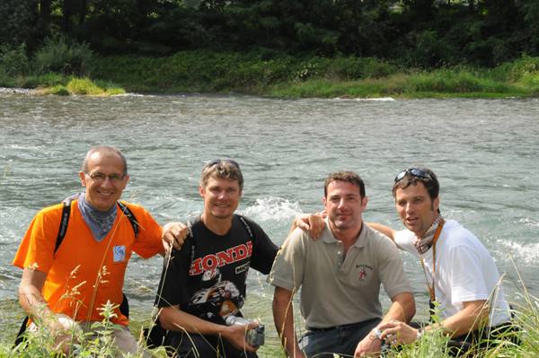 Four smiling men sit on grassy riverbank in daylight
