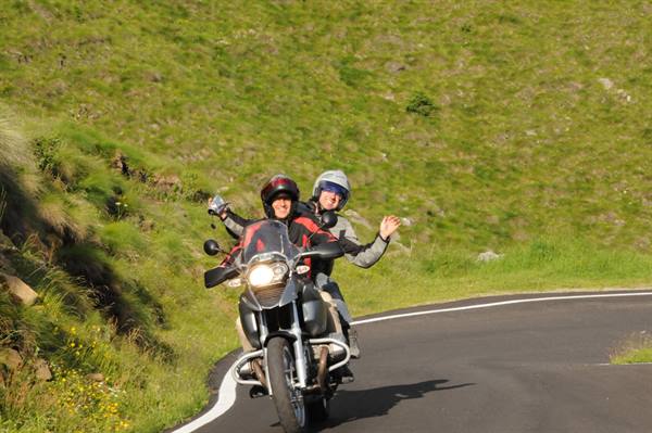 Couple waving happily from motorcycle on curvy road amid green hills