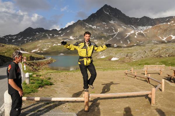 Motorcyclist in yellow suit jumps on a wooden fence, alpine lake and snowy peaks