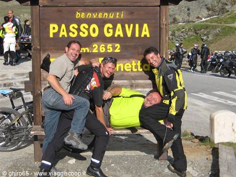 Four motorcyclists laughing in front of the wooden Passo Gavia sign