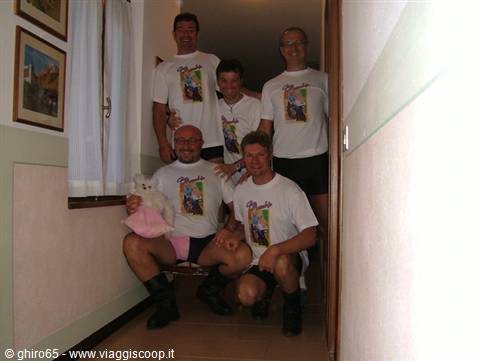 Five men in white t-shirts posing in a narrow hallway, one holding a plush toy