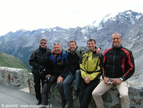 Six motorcyclists in jackets posing on a stone wall, snowy peaks behind