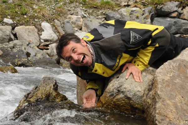 Man in yellow motorcycle jacket leans on rock touching mountain stream water