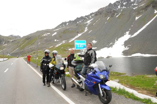 Four motorcyclists in gear beside two bikes at Picco della Mirandola pass, lake and snow