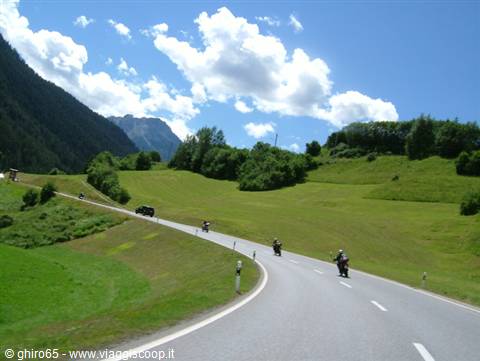 Motorcyclists riding on asphalt road between green meadows and mountains under white clouds