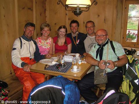 Six smiling biker friends around a wooden table in an alpine mountain hut