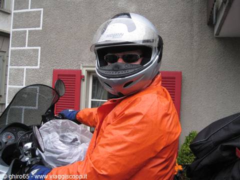 Motorcyclist in silver helmet and orange jacket in front of a house with red shutters