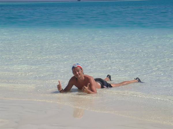Guy lying in shallow turquoise water giving thumbs up on white sand