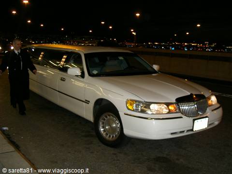 White stretch limousine parked at night, chauffeur in black jacket nearby, city lights behind
