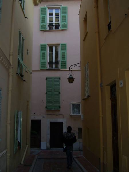 Person in a narrow alley between pastel buildings with green shutters and lantern