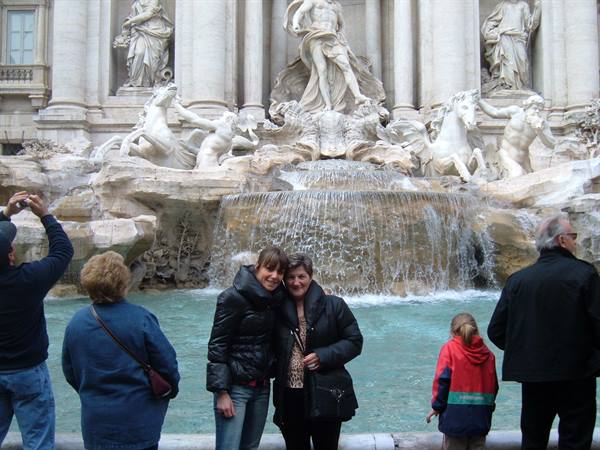 Two smiling tourists pose before Rome's Trevi Fountain, water cascading among marble horses