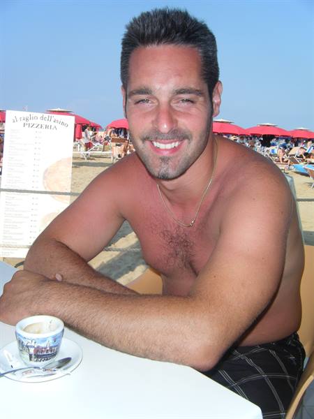 Shirtless man smiling at a beach café table, red umbrellas behind