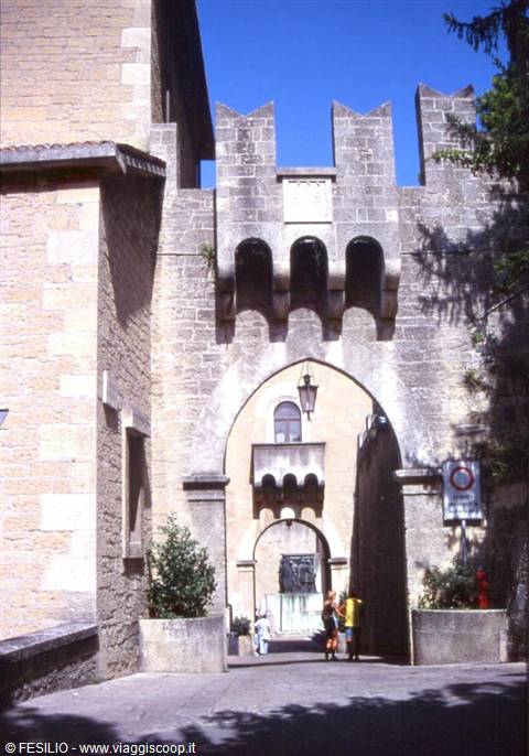 Stone gate with battlements and pointed arches, two people walking beneath