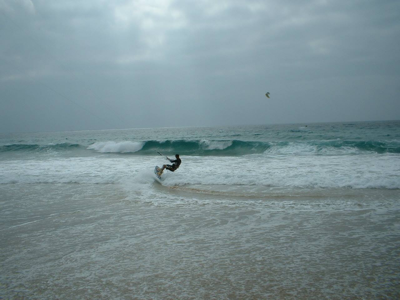 Kitesurfer riding turquoise waves, grey sky, foam on the shore