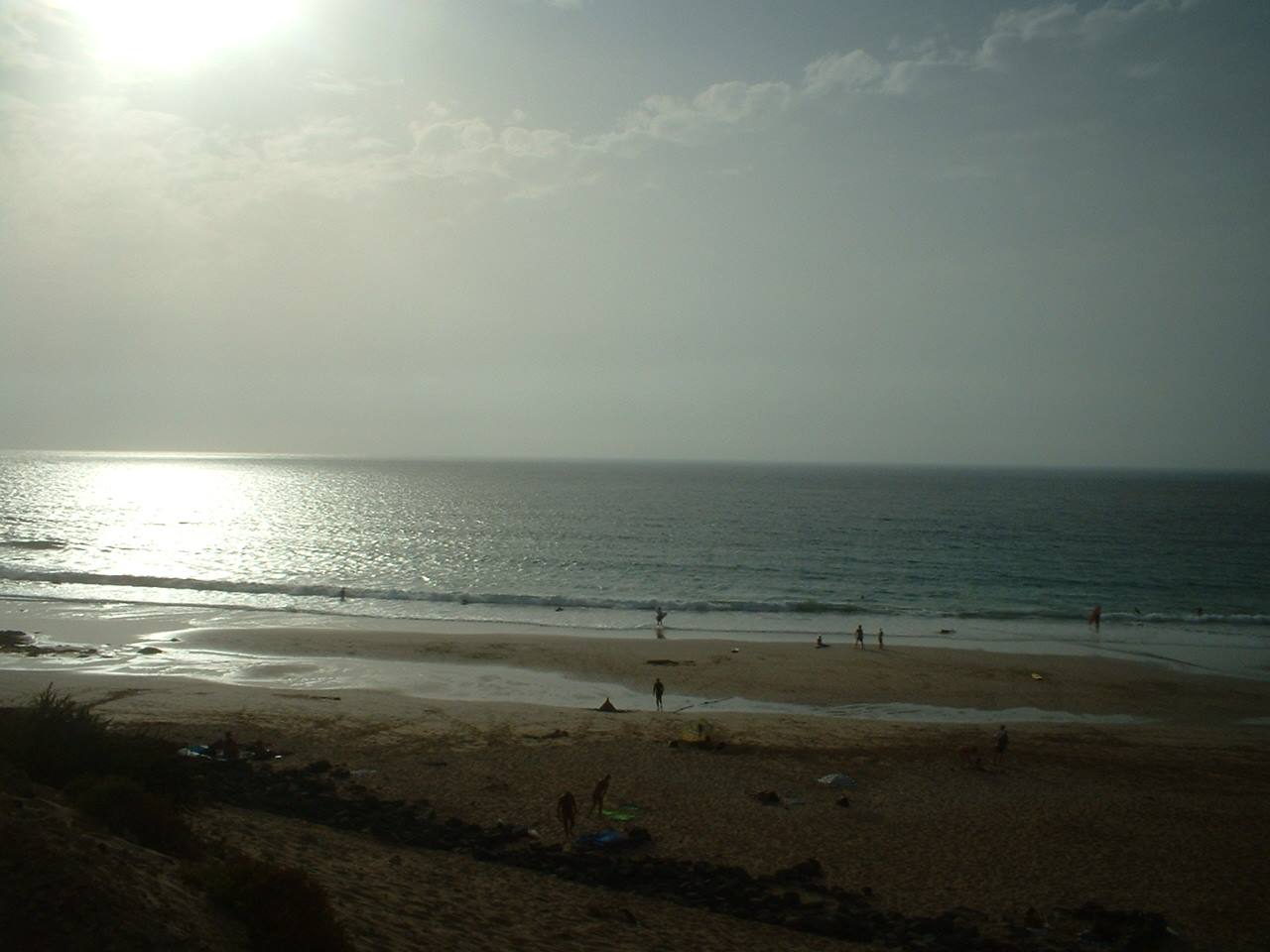 Hazy sun over dark sandy beach with people in the waves