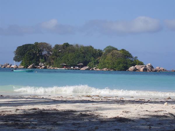 Tropical island with trees and parasols among granite rocks, waves on the beach