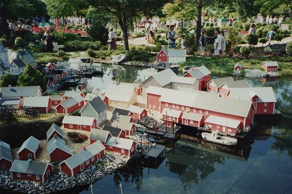Miniature village with red wooden houses by a lake, visitors strolling among gardens