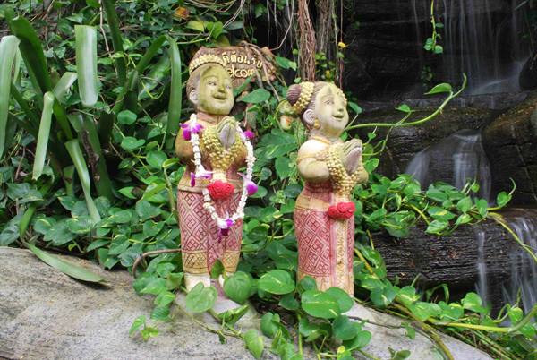Stone statues with joined hands and flower garlands among plants and waterfall