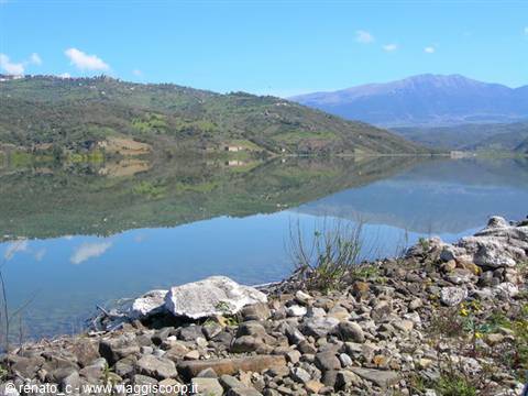 Lake surrounded by green hills and blue mountains, rocky shore in foreground