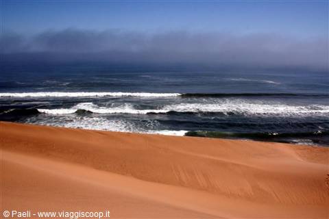 Reddish sand dunes plunge into ocean waves under coastal fog