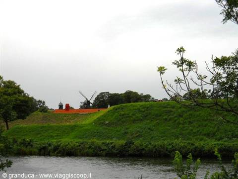 Windmill behind grassy rampart across a moat, leafy branches in foreground