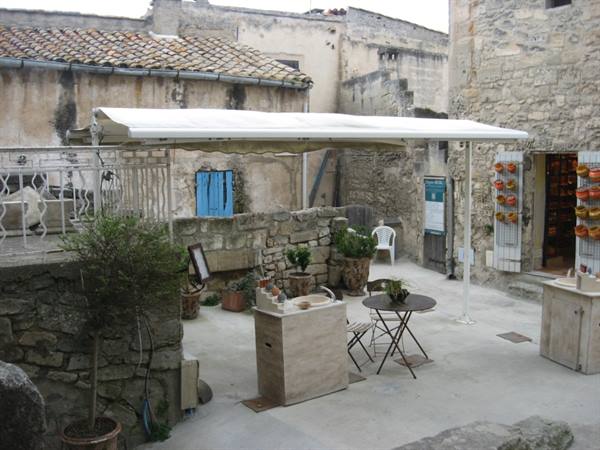 Stone courtyard with white awning, small table and pots among rustic houses, Les Baux