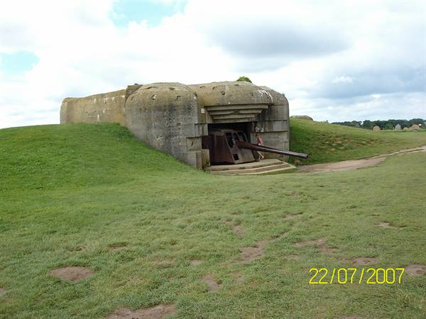 Concrete bunker with protruding German gun, green grass field, cloudy sky, Longues-sur-Mer