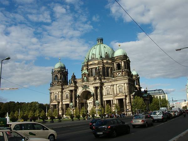 Berlin Cathedral stone facade with green domes, traffic passing under cloudy sky