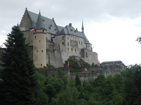 Vianden Castle rises on a rocky cliff among trees, with flags and a grey sky
