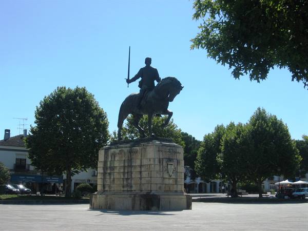 Bronze equestrian statue with raised sword on stone base, trees and white buildings under blue sky