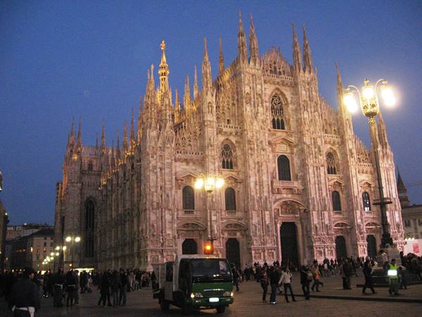 Milan Cathedral lit at dusk, tourists and a van on the square