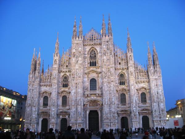 Milan Cathedral, white marble facade lit at dusk, crowd in the square