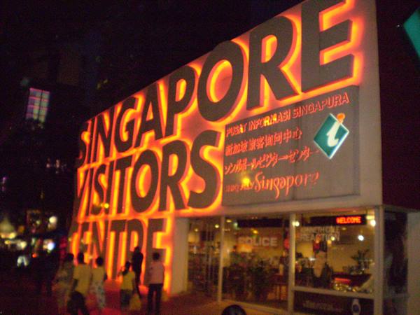 Illuminated Singapore Visitors Centre sign at night with pedestrians walking by
