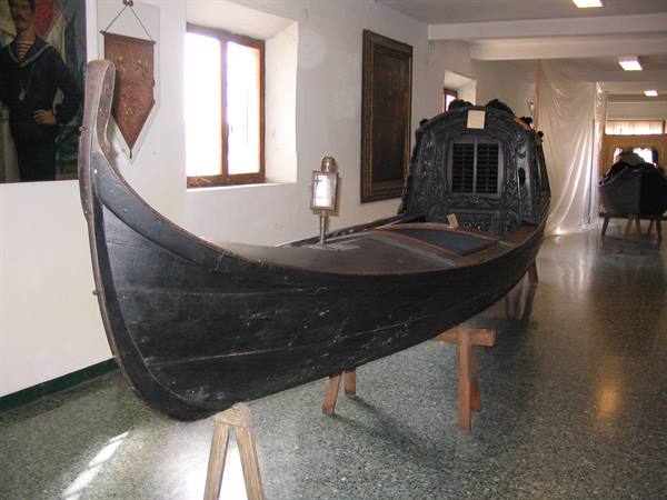 Antique black gondola displayed on wooden trestles inside a naval museum, daylight from windows