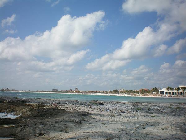 Grey rocks by sandy beach with hotels and palms under cloudy sky