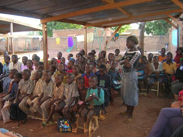 Woman with crossed arms speaks to children seated under a wooden shelter