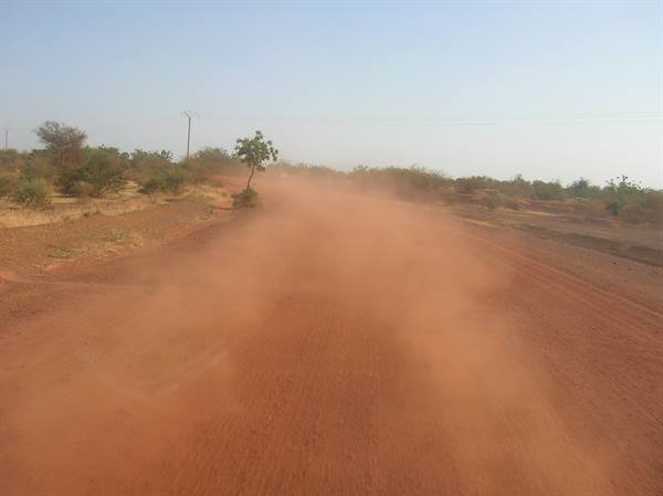 Red dust rising on a dirt road among dry bushes and scattered trees