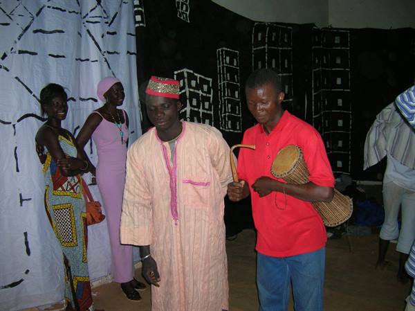 Men in boubou robes and talking drum before painted backdrops, traditional theater